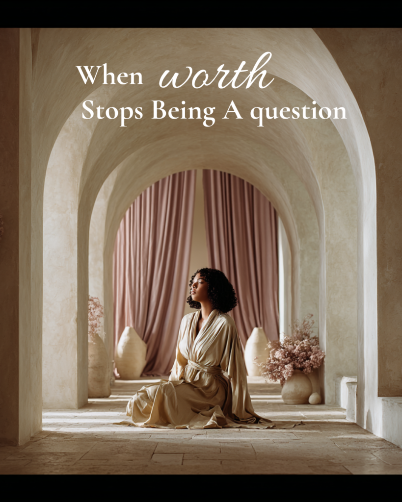 A woman seated calmly beneath soft stone arches and blush curtains, embodying embodied self-worth and quiet feminine authority as worth stops being a question.