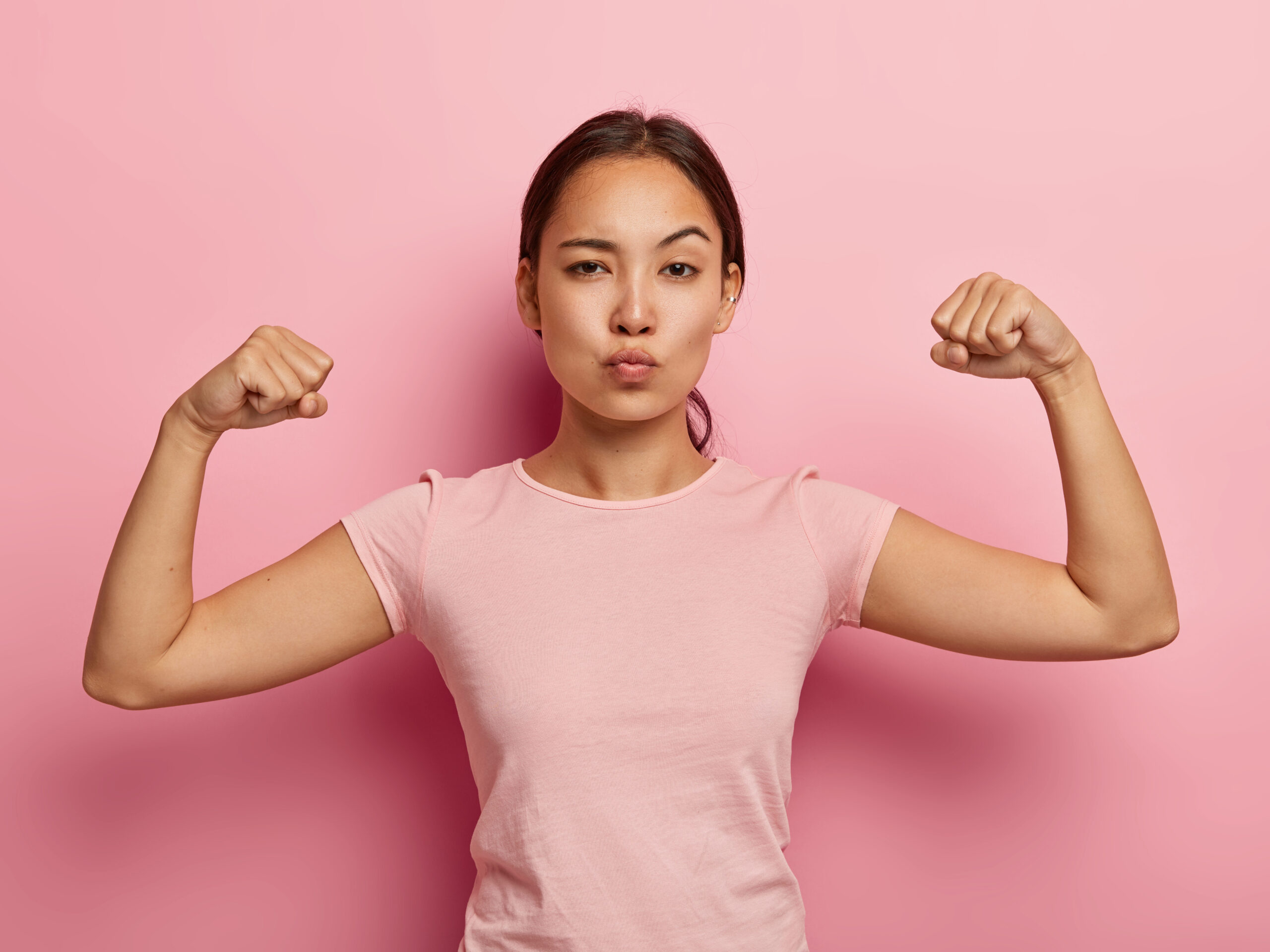 Confident young woman flexing her muscles with a determined expression against a pink background, symbolizing personal empowerment and strength. Photo of self confident serious Asian girl keeps lips folded, shows her muscle and strength, wears no make up, dressed in casual t shirt, isolated on pink wall with copy space above for information