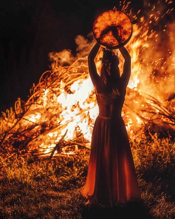 A striking, dramatic scene featuring a woman standing before a raging fire at night. She wears a long skirt and a crown-like headpiece, lifting a round drum above her head. The flames create an intense, powerful energy, symbolizing ritual, transformation, or divine feminine power.