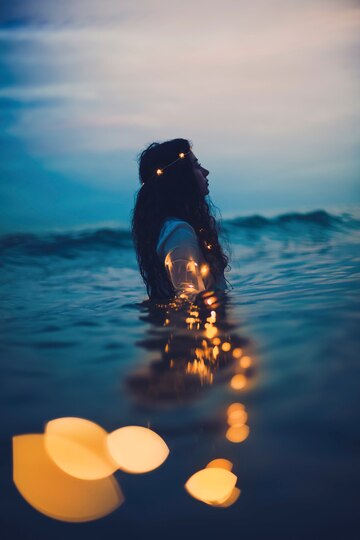 The image features a woman partially submerged in water during twilight or early evening. She has long, flowing hair and wears a delicate headband adorned with small glowing lights. Her arm is extended backward, also adorned with glowing fairy lights, creating a striking reflection on the water’s surface. The background consists of a calm sea or lake with soft waves, under a sky with a gradient of blue and purple hues. In the foreground, a few blurred golden lights float on the water, adding a dreamy, mystical atmosphere to the scene. The overall composition evokes a sense of serenity, magic, and ethereal beauty.
