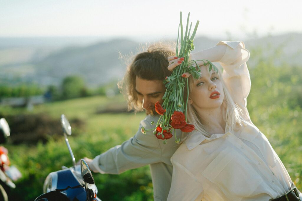 A couple poses with flowers on a moped, surrounded by a scenic landscape representing feminine energy and healthy masculinity