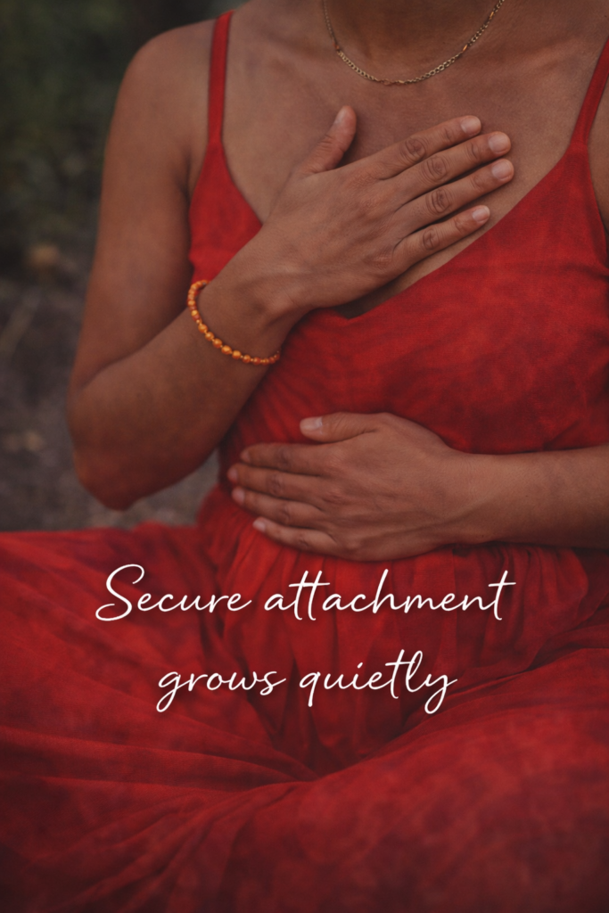 A woman in a red dress sits peacefully with one hand on her heart and the other on her belly, symbolizing grounding and self-soothing, with the words “Secure attachment grows quietly” written across the image.