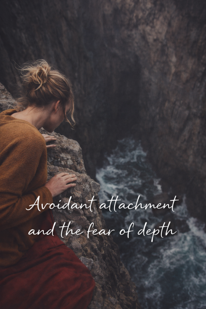 A woman sits on the edge of a rocky cliff, leaning forward as she looks down at the dark ocean below. She wears a cozy mustard sweater and has her hair in a loose bun, capturing the feeling of distance, reflection, and emotional hesitation.