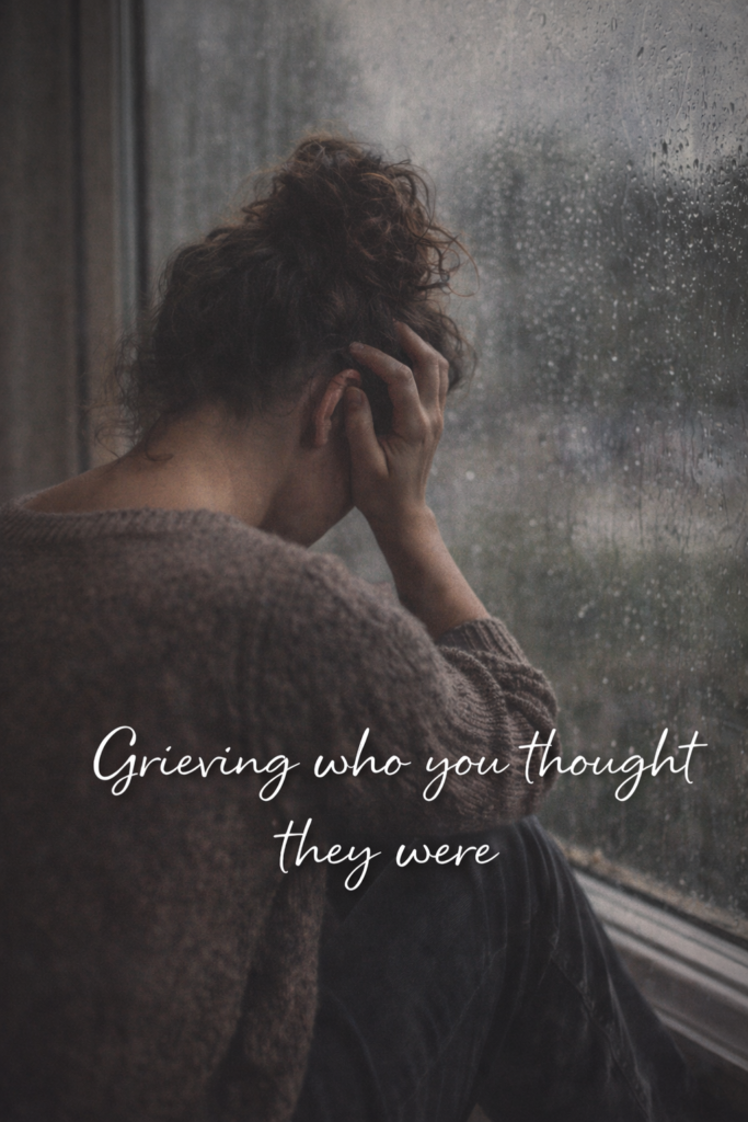 A woman sits by a rain-streaked window with her head in her hand, wearing a cozy brown sweater. Her hair is tied in a messy bun, and the scene feels quiet, reflective, and emotional, capturing the mood of grief and introspection.
