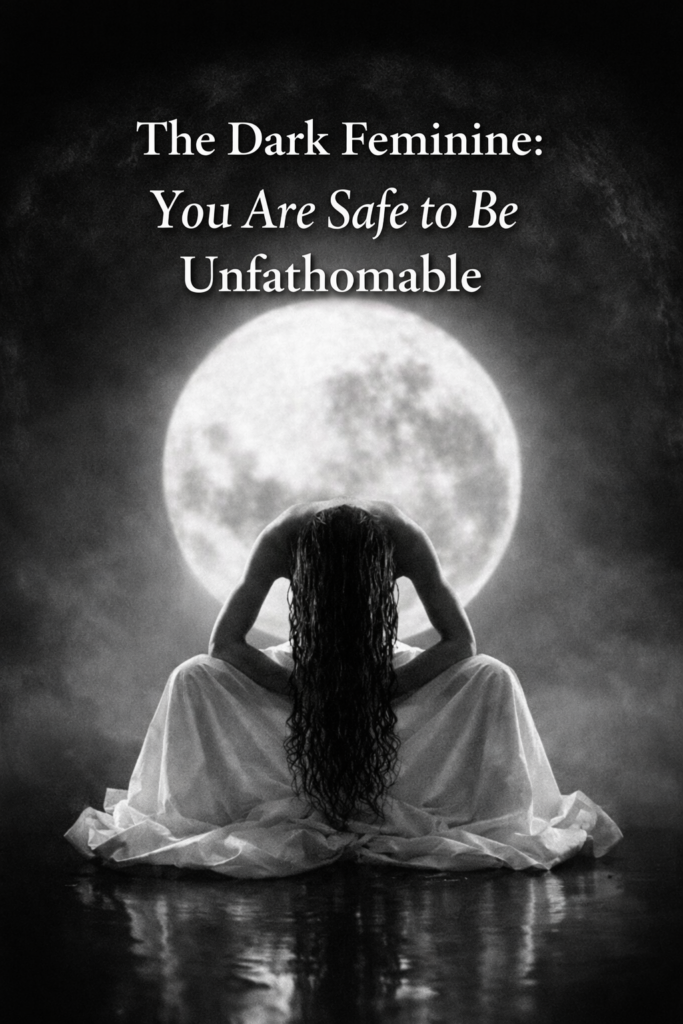 Black and white image of a woman seated with her back to the viewer, long dark hair cascading forward, wrapped in a flowing white garment, silhouetted against a large full moon. Soft mist surrounds her, creating a womb-like, protective atmosphere. Text overlay reads: “The Dark Feminine: You Are Safe to Be Unfathomable.”