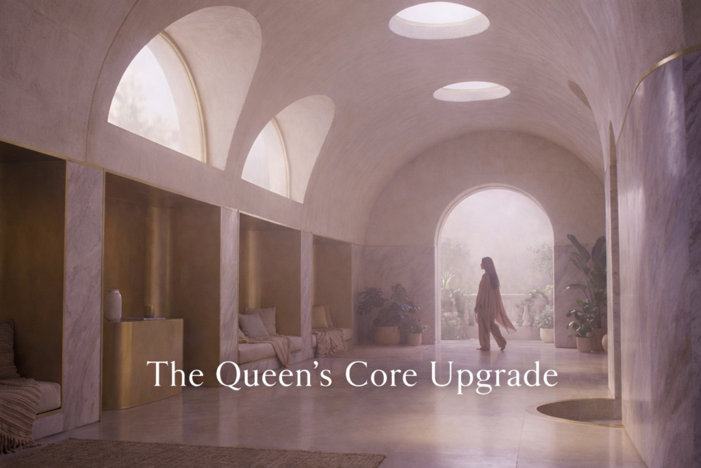 Serene, softly lit interior sanctuary in blush-pink and warm stone tones, with arched ceilings and gentle daylight. A distant, fully clothed feminine silhouette walks calmly through the space, conveying embodied sovereignty and quiet power, with the text overlay “The Queen’s Core Upgrade